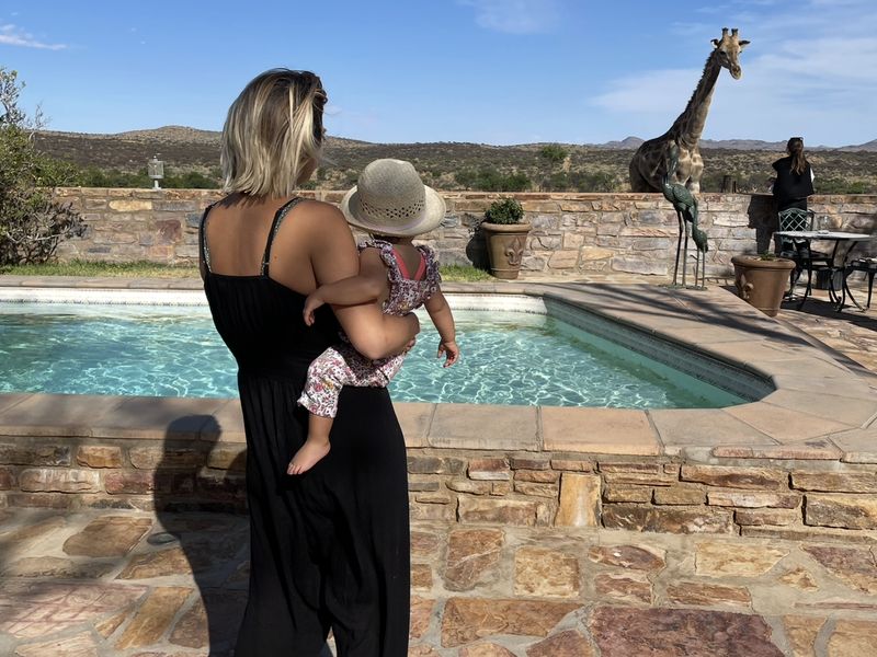 Une maman et son bébé au bord d'une piscine, face à une girafe, en Namibie