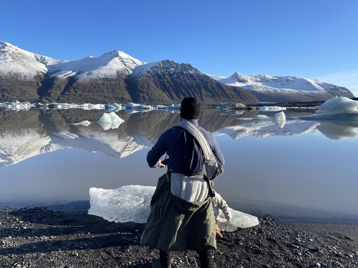 Papa et notre fille au bord d'un lagon glaciaire où flottent des icebergs, face aux montagnes enneigées